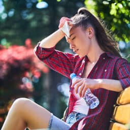 Dehydrated woman holds a water bottle and wipes her sweaty forehead on a hot, humid summer day.