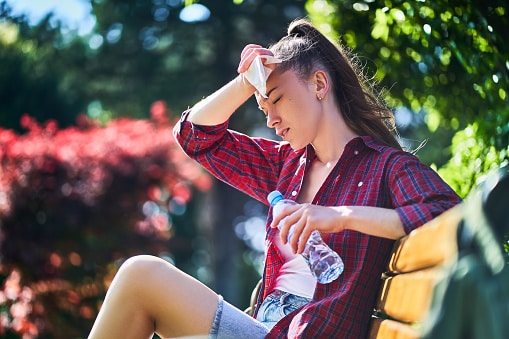 Dehydrated woman holds a water bottle and wipes her sweaty forehead on a hot, humid summer day.
