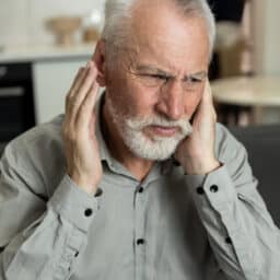 Portrait of unhappy senior man suffering from ear infection and headache at home. Irritated male with otitis covering head with hands, hearing loud noise